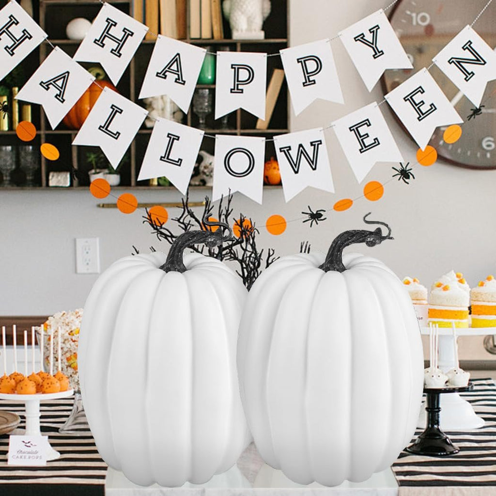 Two large white artificial foam pumpkins with black stems on a table, with a 'Happy Halloween' banner and party treats in the background.