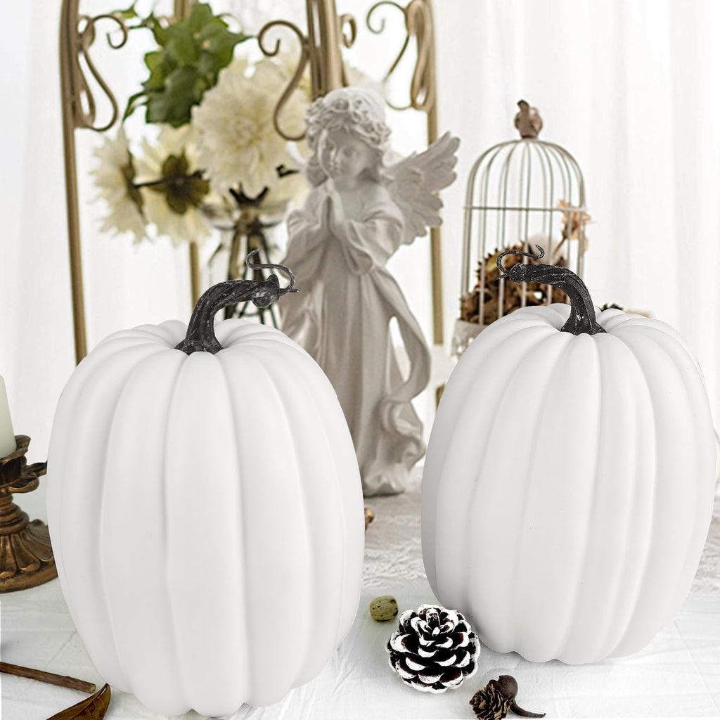 Two white artificial pumpkins with dark stems on a white tablecloth, surrounded by decorative fall elements.