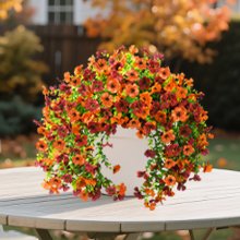 Artificial orange and brown daisy flowers with eucalyptus leaves in a white pot on a table outdoors.