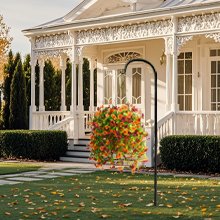 Artificial hanging flowers with eucalyptus leaves in orange and brown hues, displayed in a basket on a shepherd's hook in front of a white house.