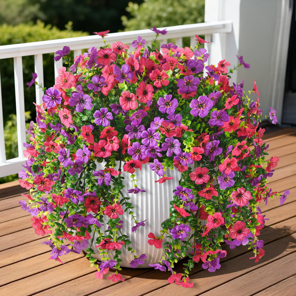 Hanging artificial flowers in a white pot on a wooden deck, featuring pink and purple blooms with green foliage.