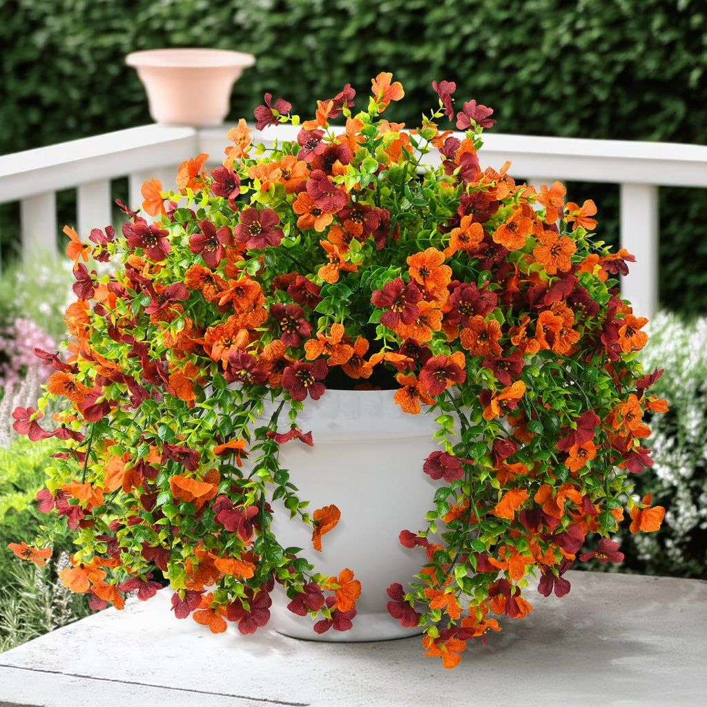 Hanging artificial flowers in orange and burgundy with green leaves in a white pot on a table