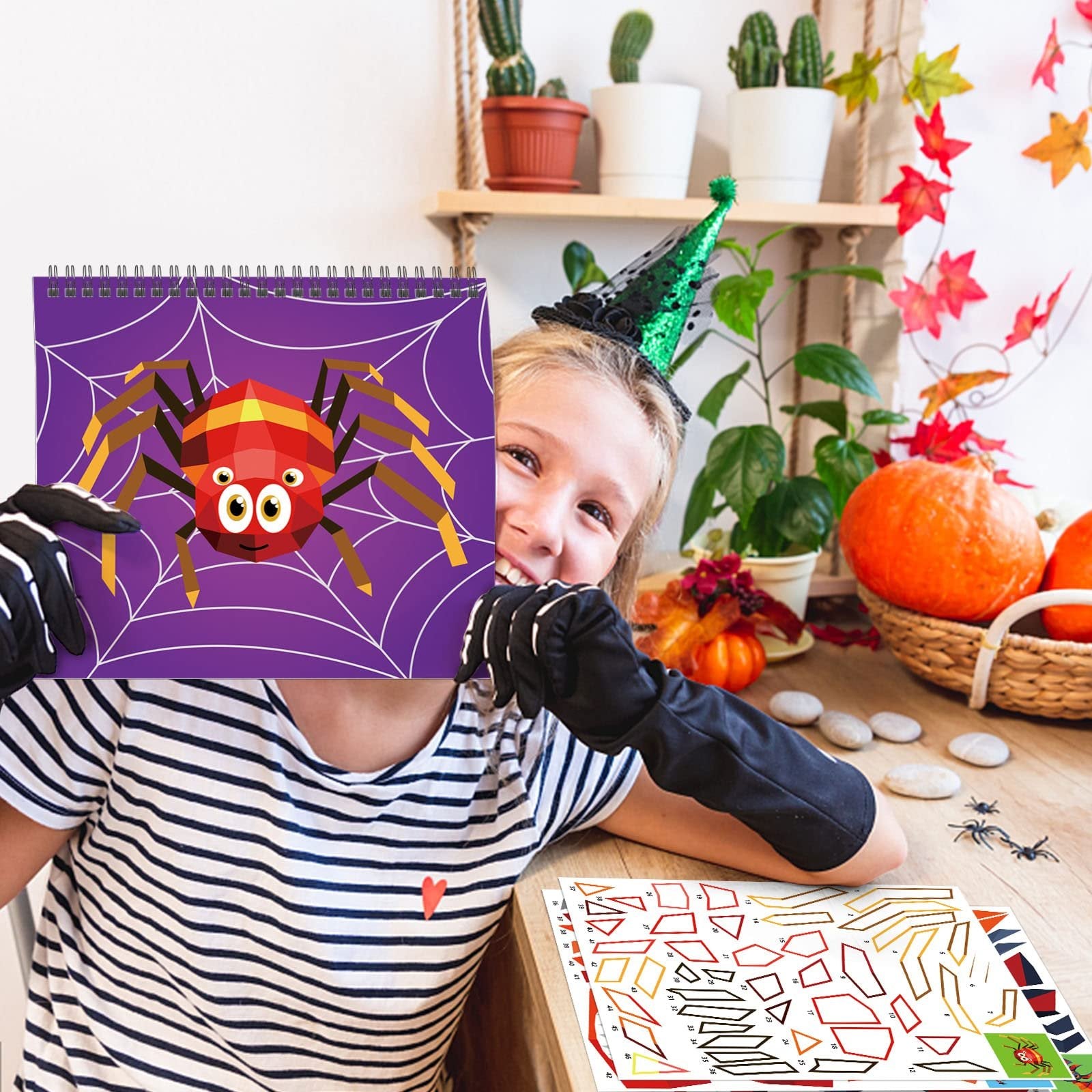 Smiling child wearing a witch hat holds up a Halloween sticker paint book featuring a spider on a web. Sticker sheets are visible on the table.