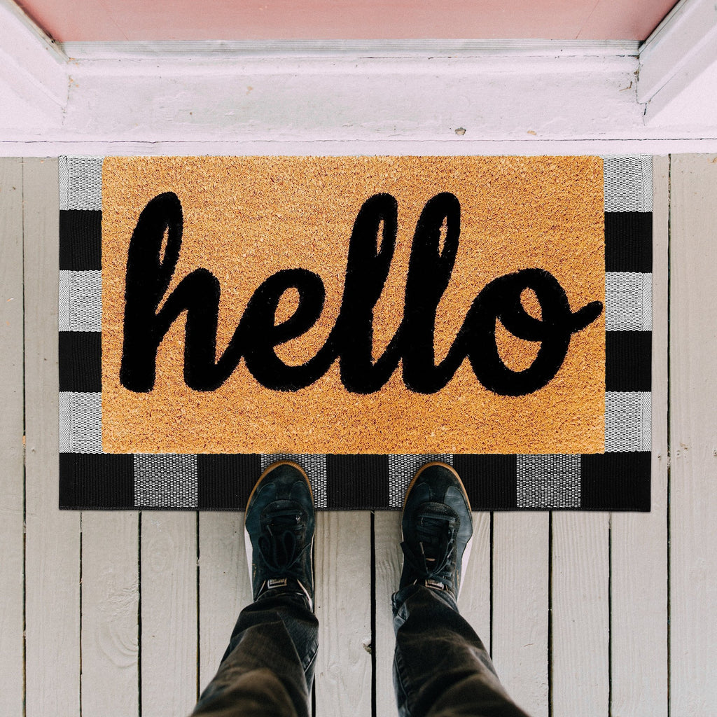 Black and white buffalo plaid doormat with 'hello' in script, shown from a first-person perspective with feet on the mat.