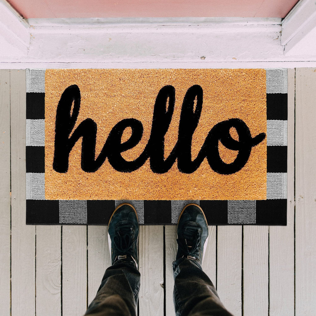 Black and white buffalo plaid doormat with "hello" in script, shown from a top-down perspective with feet wearing sneakers standing on it.