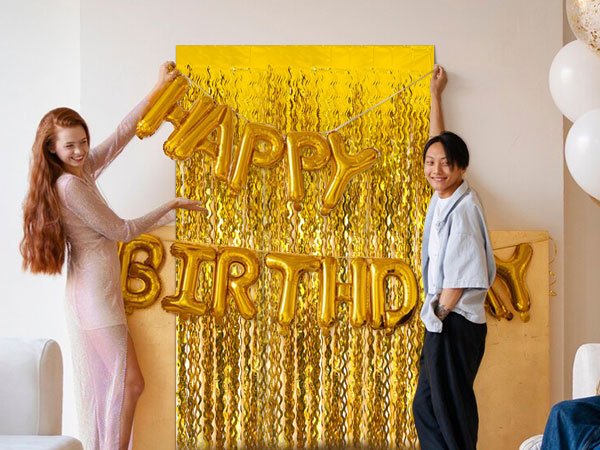 Two people setting up a gold foil fringe curtain backdrop with gold 'HAPPY BIRTHDAY' balloons for a party.