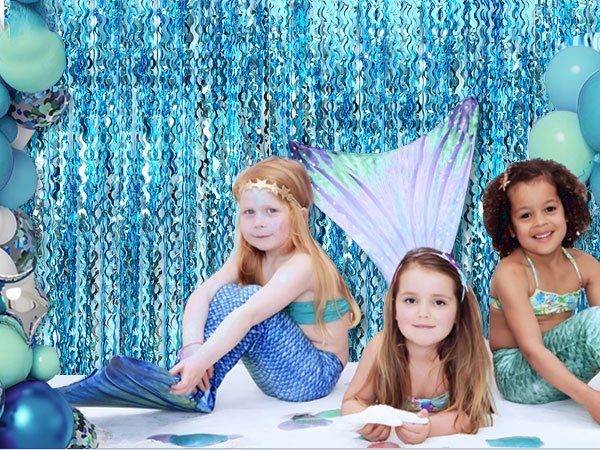 Three young girls in mermaid costumes posing in front of a pale blue foil fringe curtain backdrop with balloons.
