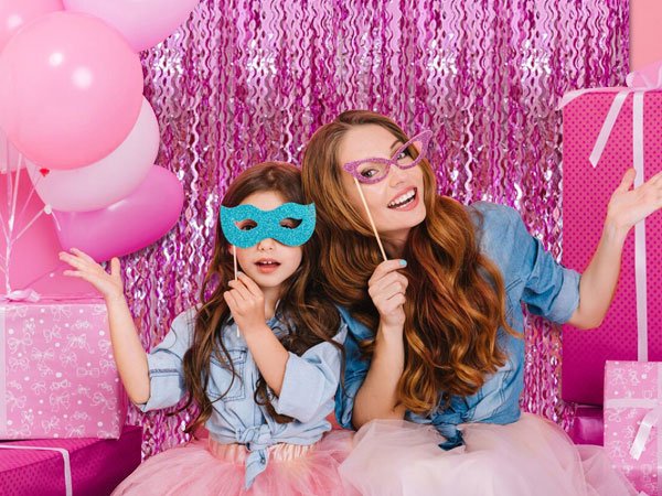 Woman and child posing with party masks in front of a pink foil fringe curtain backdrop with balloons and gifts
