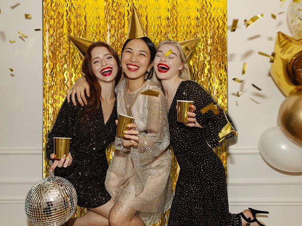 Three women celebrating with gold cups and confetti, with a gold fringe backdrop and balloons.