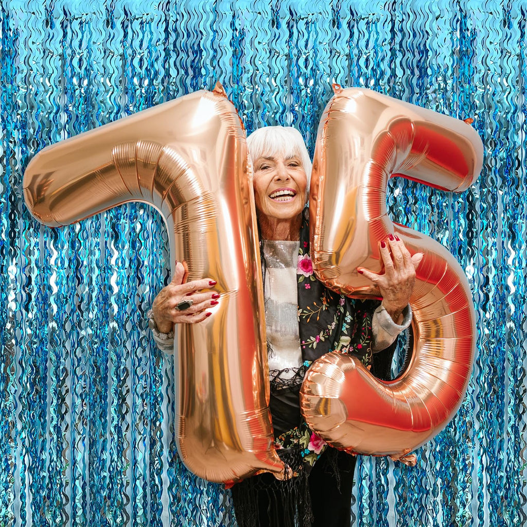 Elderly woman holding rose gold number balloons '75' in front of a blue tinsel fringe backdrop