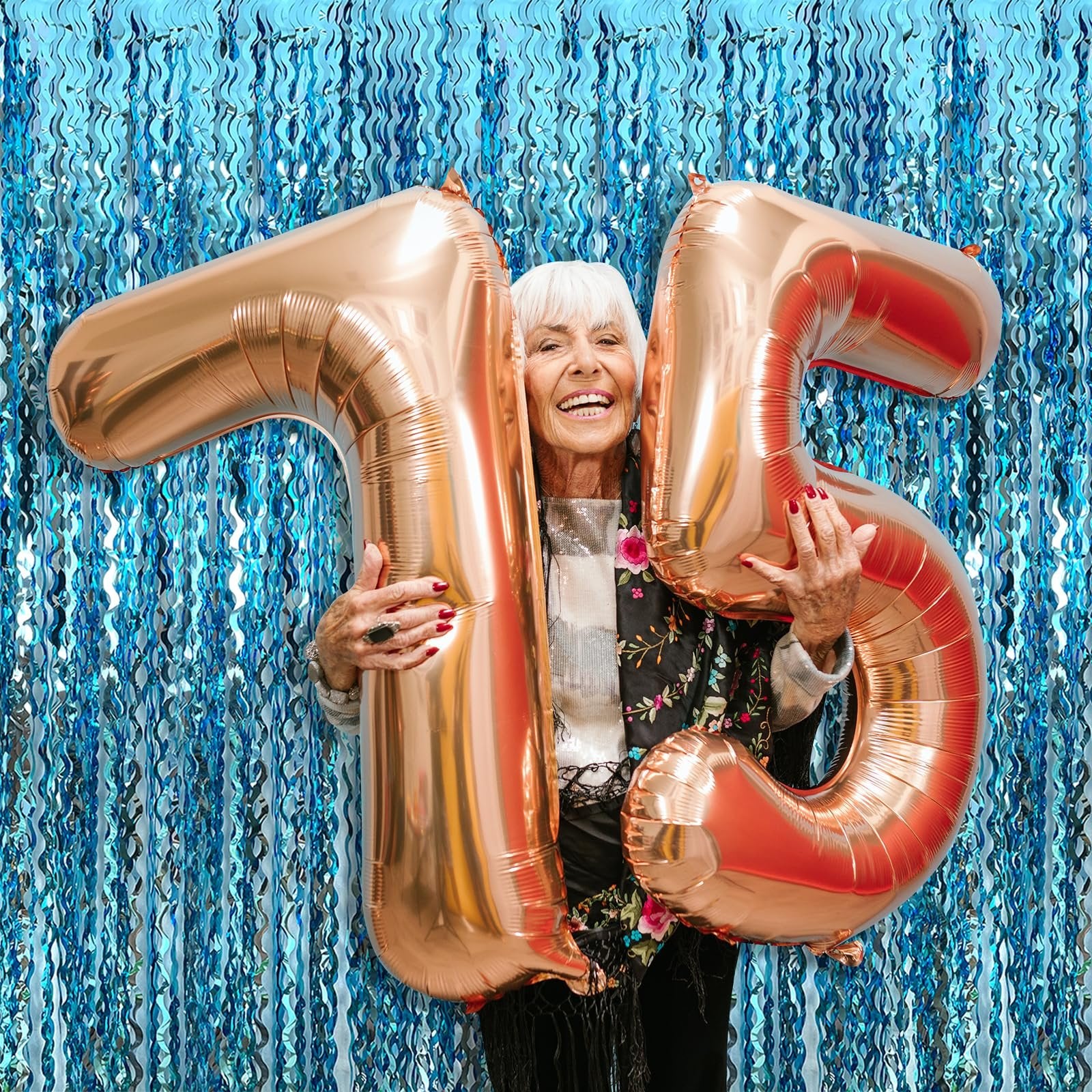 Elderly woman holding rose gold number balloons '75' in front of a blue tinsel fringe backdrop