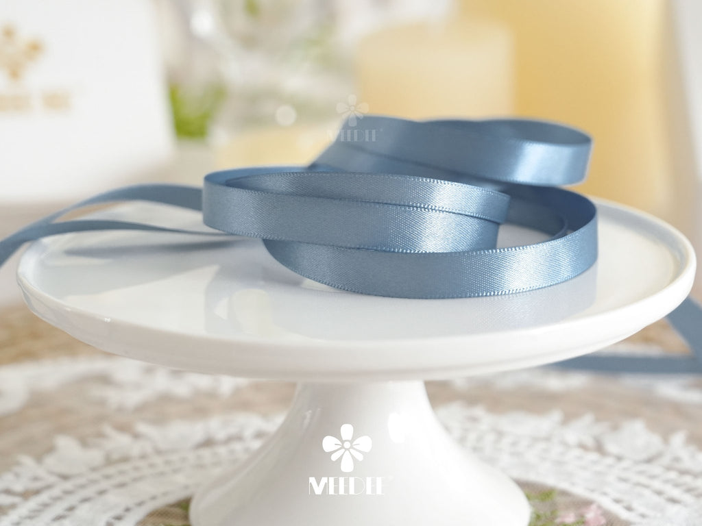 Dusty blue double sided satin ribbon coiled on a white cake stand with blurred candles in the background.