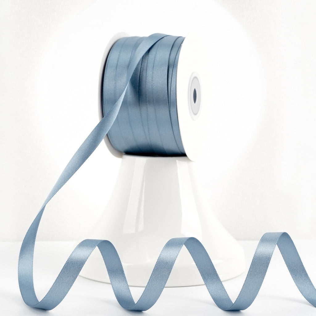Spool of dusty blue double-sided satin ribbon with a length of ribbon unspooled in a wavy pattern.