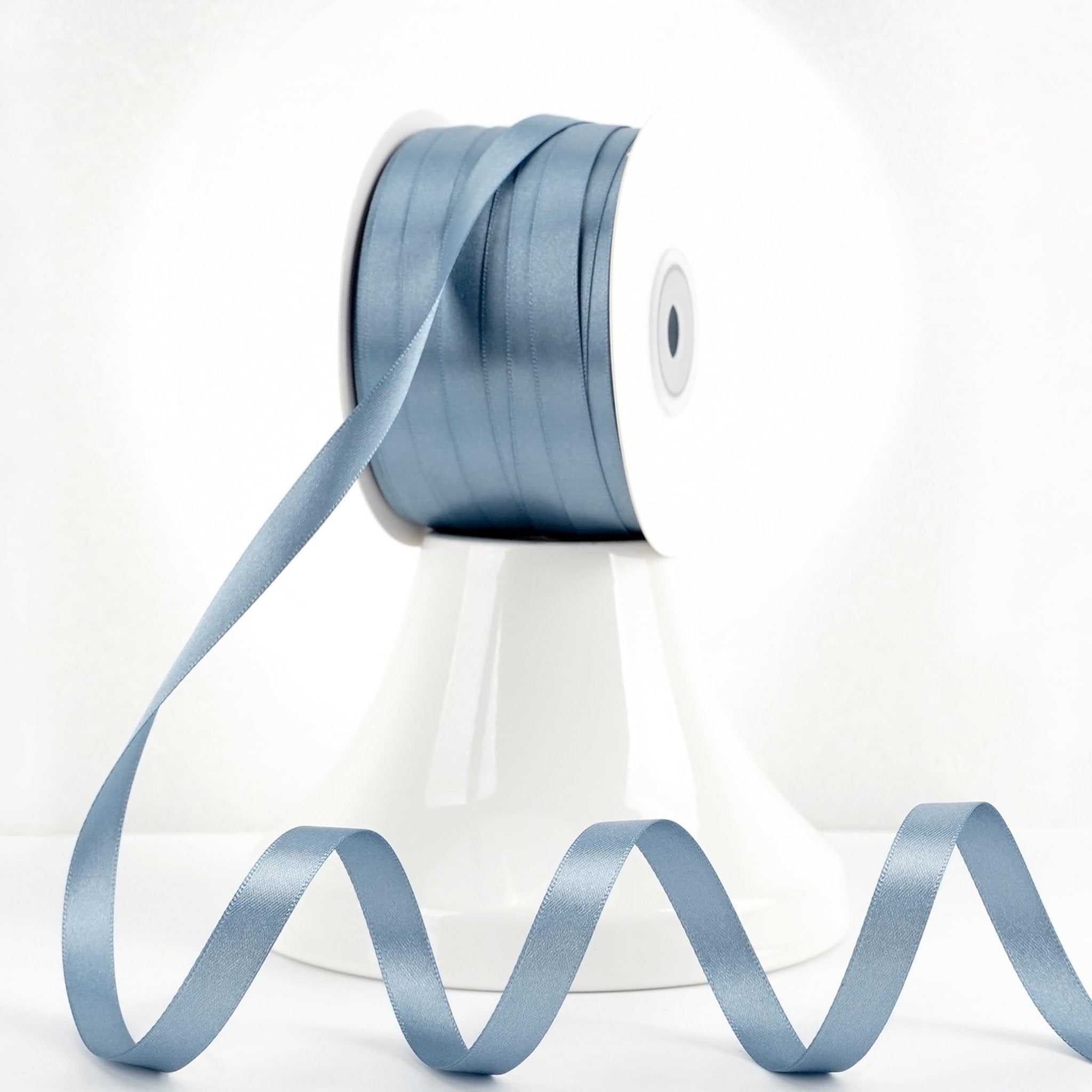 Spool of dusty blue double-sided satin ribbon with a length of ribbon unspooled in a wavy pattern.