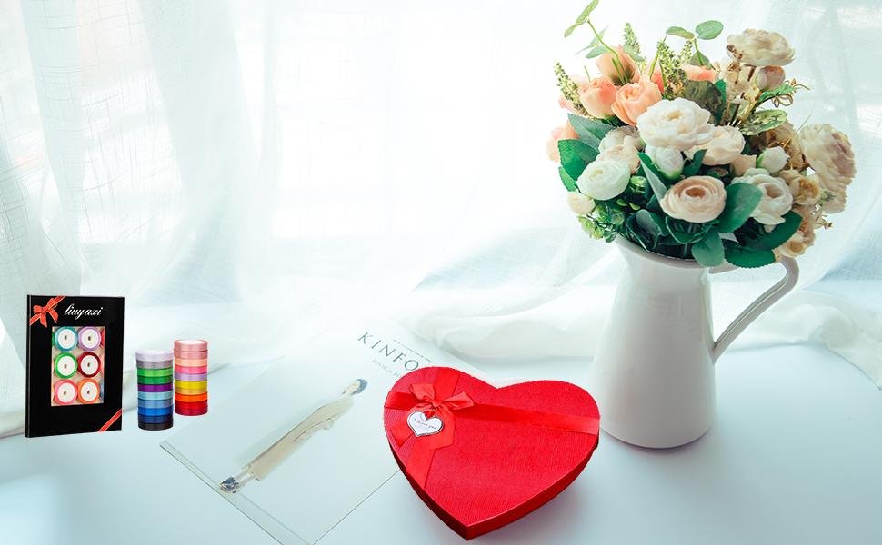 Set of 20 colorful satin ribbon rolls in a box and stacked, next to a heart-shaped gift box and flowers.