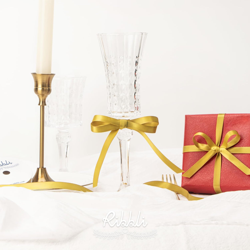 Gold ribbon tied in a bow around a champagne flute next to a gift-wrapped box and candlestick.