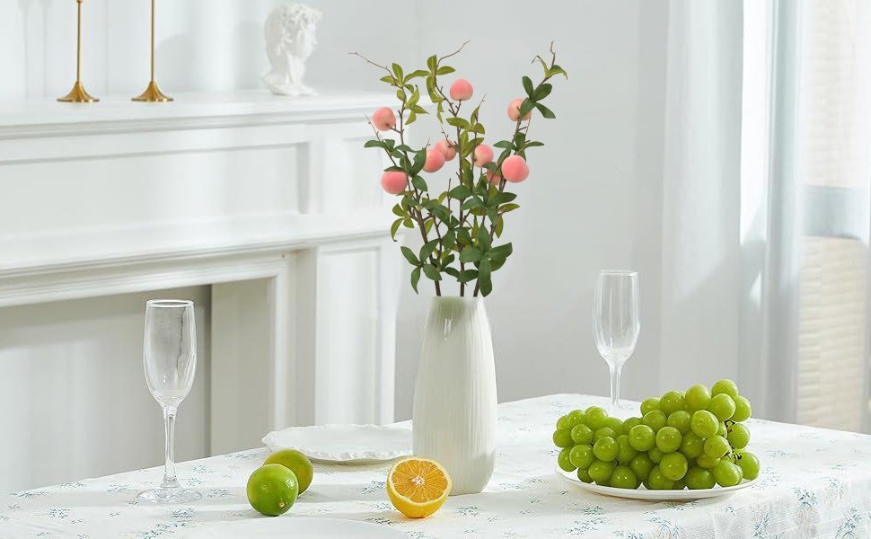 Artificial peach branches with pink fruit and green leaves in a white vase on a table with grapes and champagne flutes.