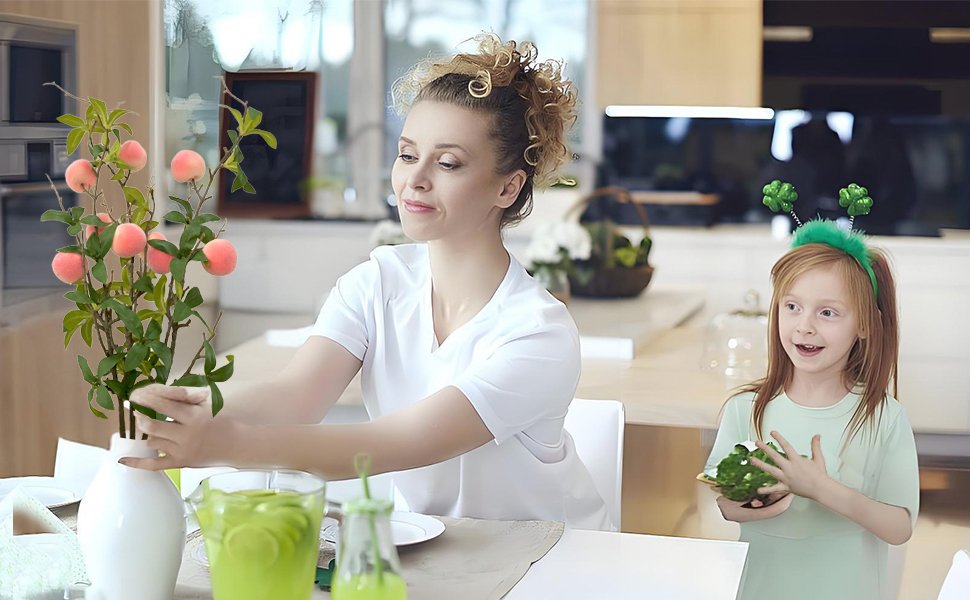 Woman arranging artificial peach branches in a vase on a table with a child holding a green item