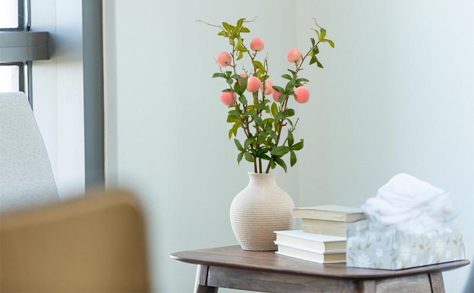 Artificial peach branches with pink fruit and green leaves in a vase on a table with books and tissues