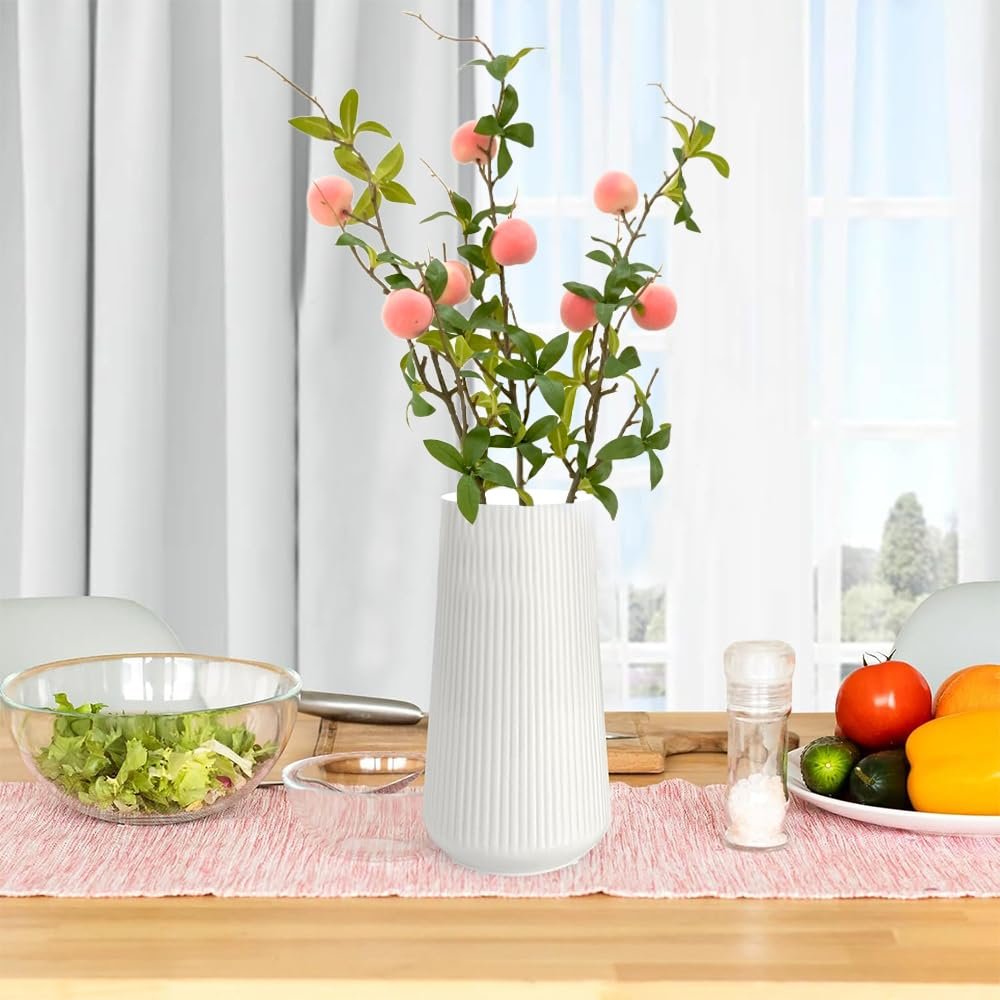 Artificial peach branches with pink fruit and green leaves in a white vase on a table with a salad bowl and vegetables.