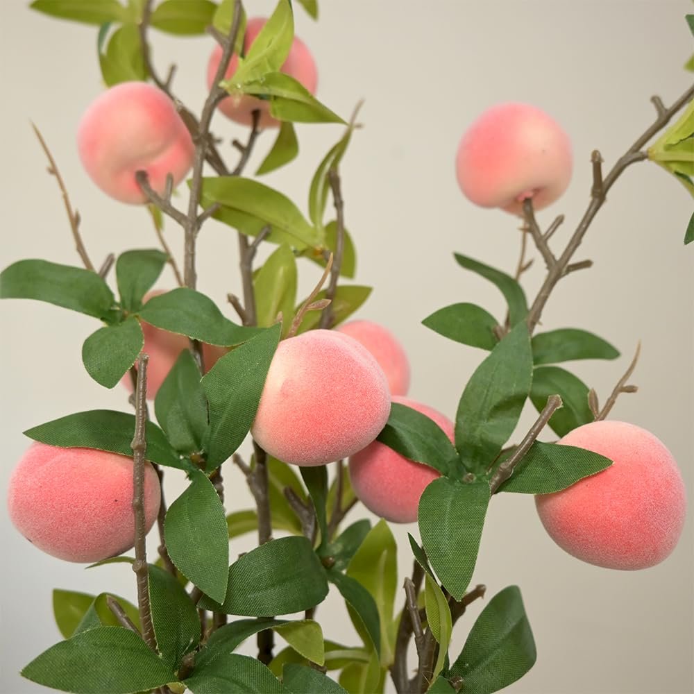 Close-up of artificial peach branches with pink fuzzy peaches and green leaves against a plain background.