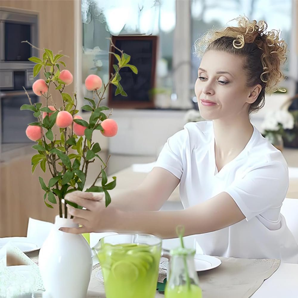 Woman arranging artificial peach branches with pink fruit and green leaves in a white vase on a table.