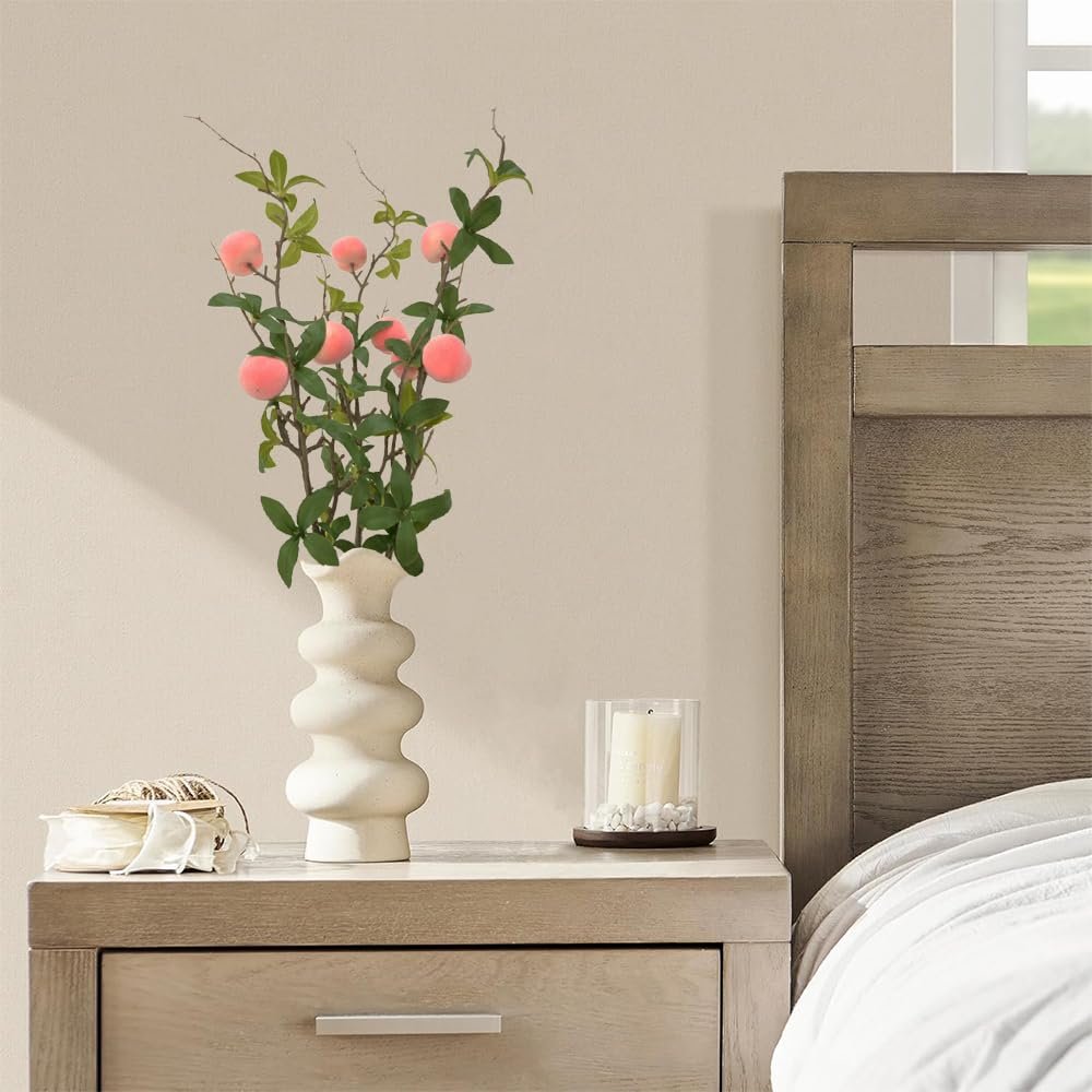 Artificial peach branches with pink fruit and green leaves in a white vase on a nightstand next to a bed.