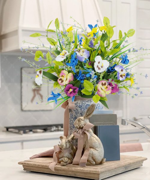 Artificial mixed pansy and wildflower bouquet in a blue and white patterned vase with two gold bunny figurines on a wooden tray.