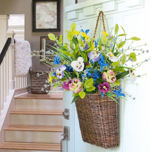 Wicker basket filled with artificial pansies and wildflowers hanging on a door, with stairs and a hallway in the background.