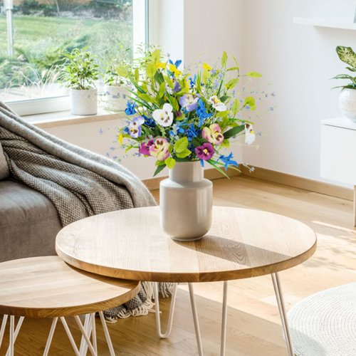 Colorful artificial pansy and wildflower bouquet in a vase on a wooden coffee table in a living room.