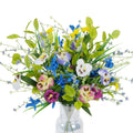 Mixed artificial pansy and wildflower bouquet in a clear glass vase, featuring purple, pink, yellow, blue, and white flowers with green foliage.
