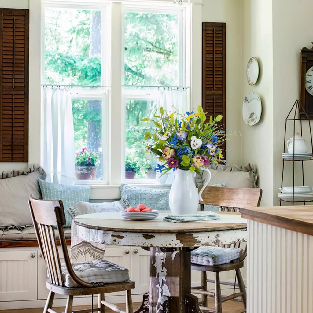 Mixed pansy and wildflower bouquet in a white pitcher on a rustic table, with chairs and a window view.