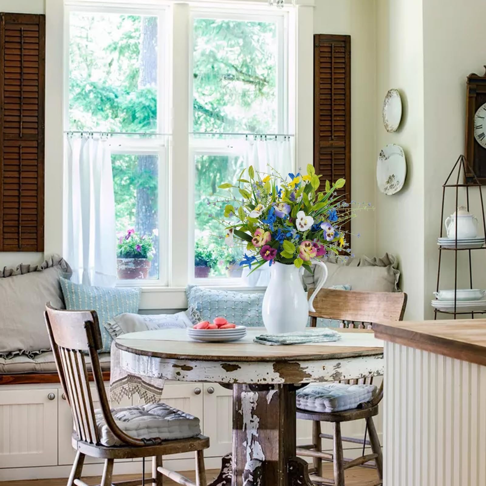 Mixed pansy and wildflower bouquet in a white pitcher on a rustic table, with chairs and a window view.