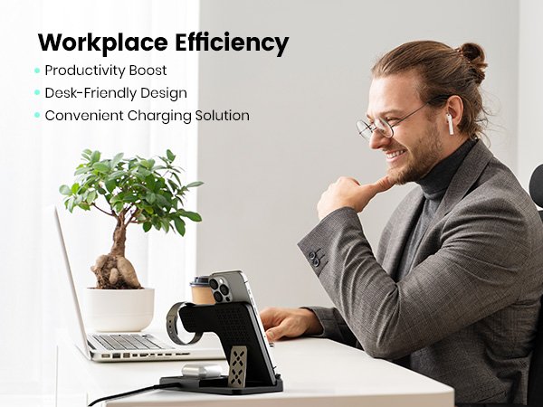 Man smiling at desk with wireless charging station holding phone, watch, and earbuds; laptop and plant nearby.