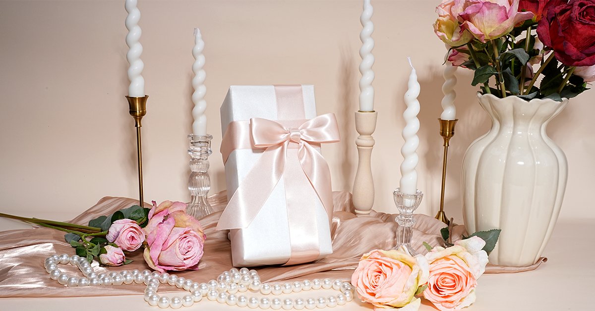 Gift-wrapped box with pink ribbon, surrounded by candles, roses, and pearls on a draped fabric.