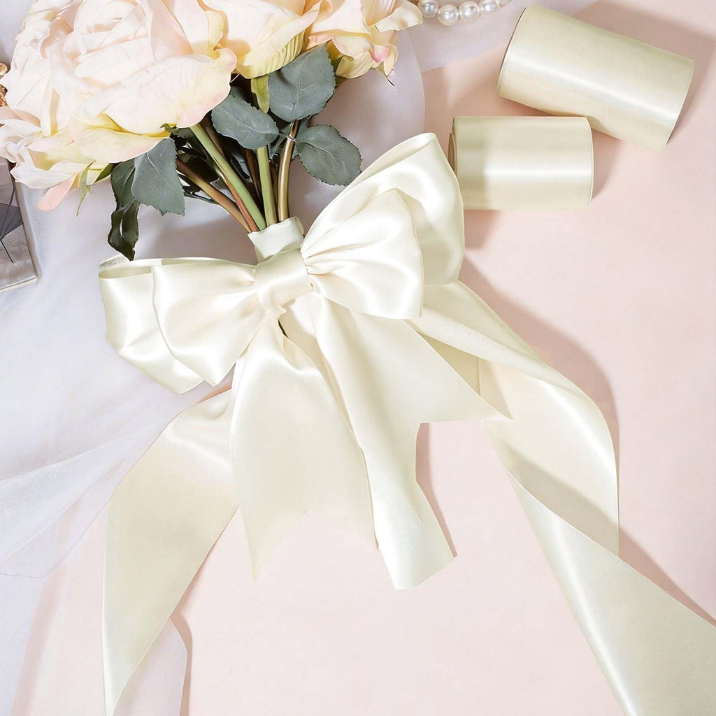 Ivory double faced satin ribbon tied in a large bow around a bouquet of pale roses, with two spools of ribbon in the background.
