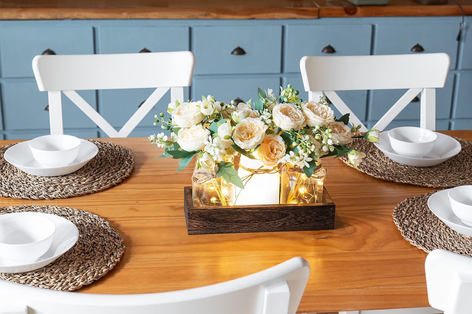 Farmhouse centerpiece with artificial flowers and fairy lights in mason jars on a wooden tray, set on a dining table.