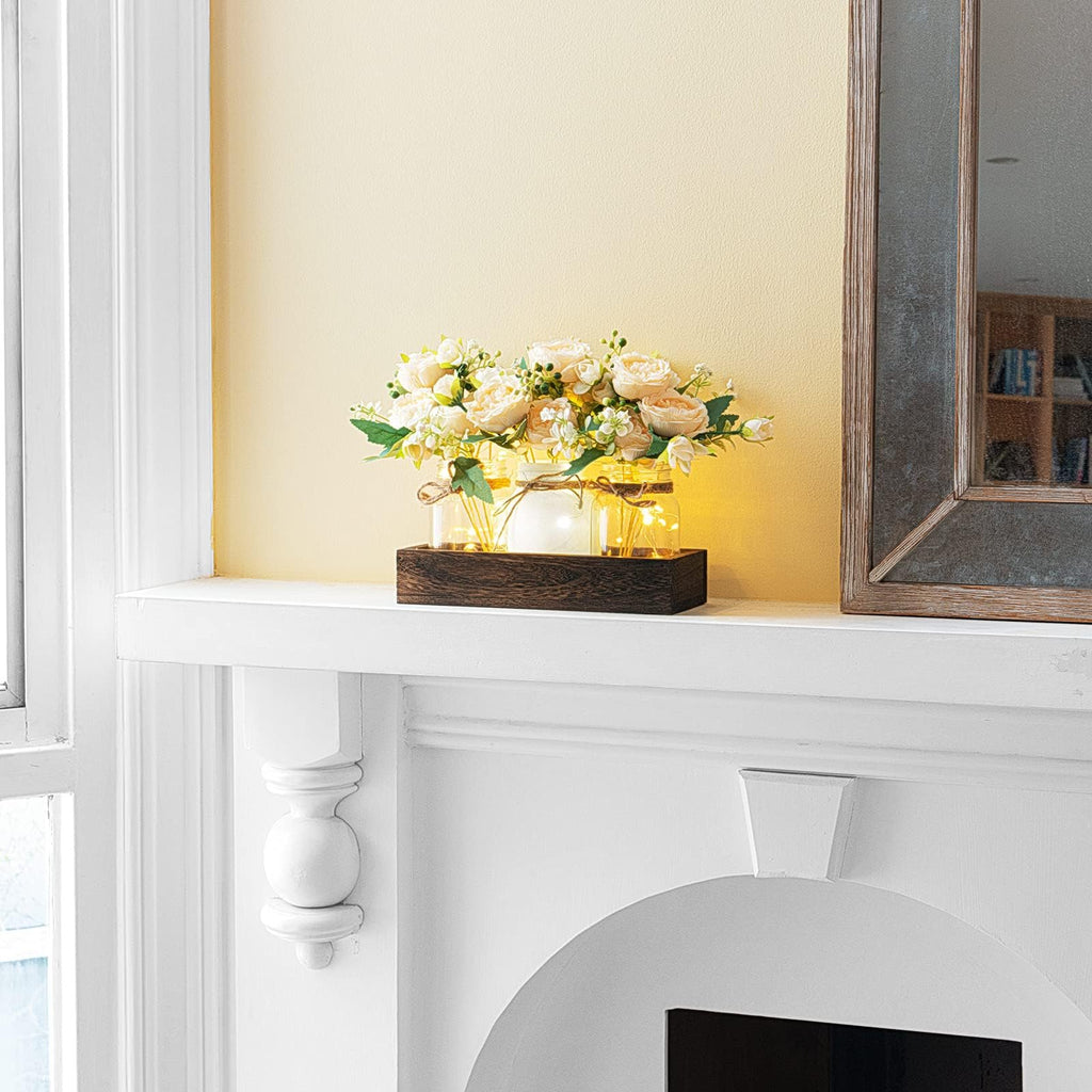 Mason jar centerpiece with artificial flowers and fairy lights on a wooden tray, displayed on a white mantelpiece.