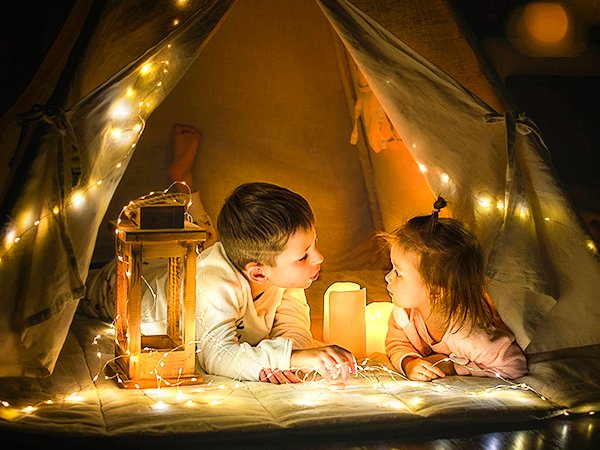 Two children lying inside a tent decorated with warm white fairy string lights and candles.
