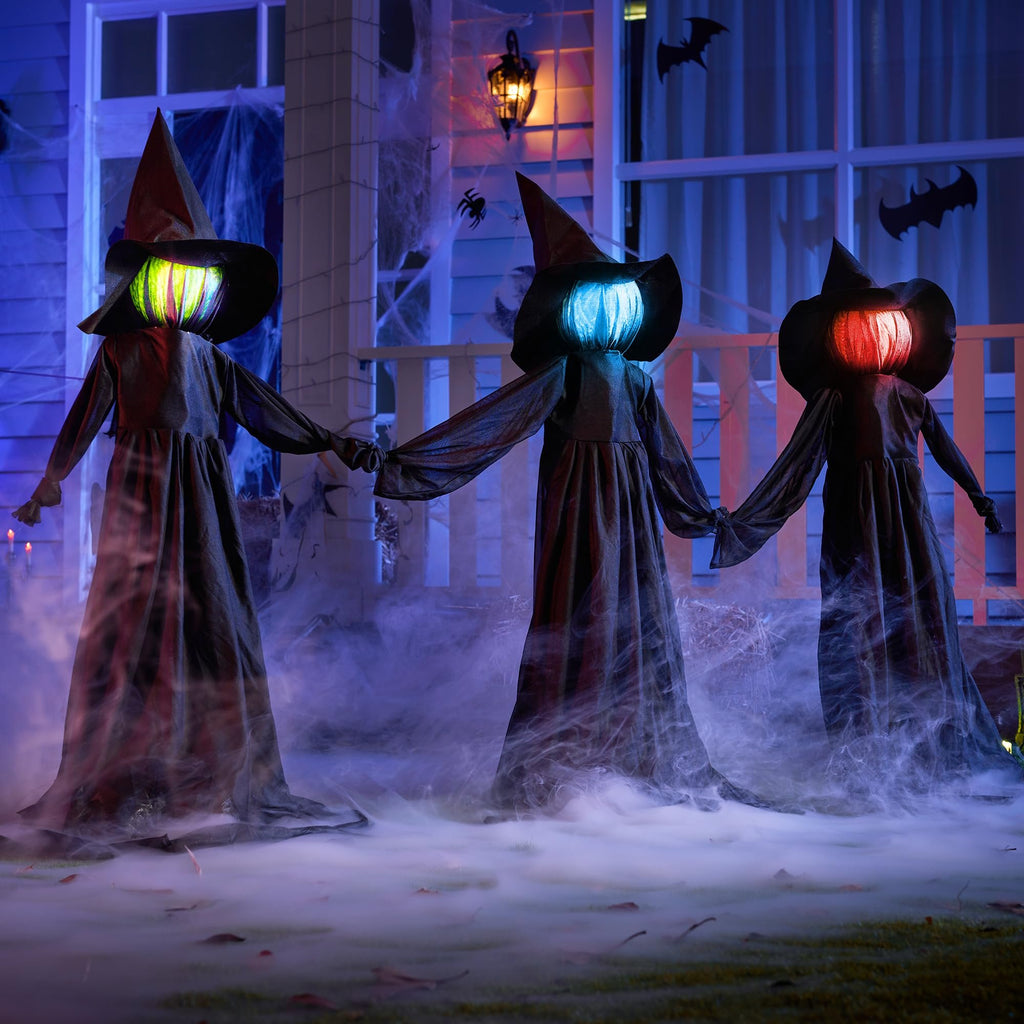 Three light-up flying witch yard stakes with flashing LED heads stand in fog on a lawn at night, decorated with spiderwebs and bats.