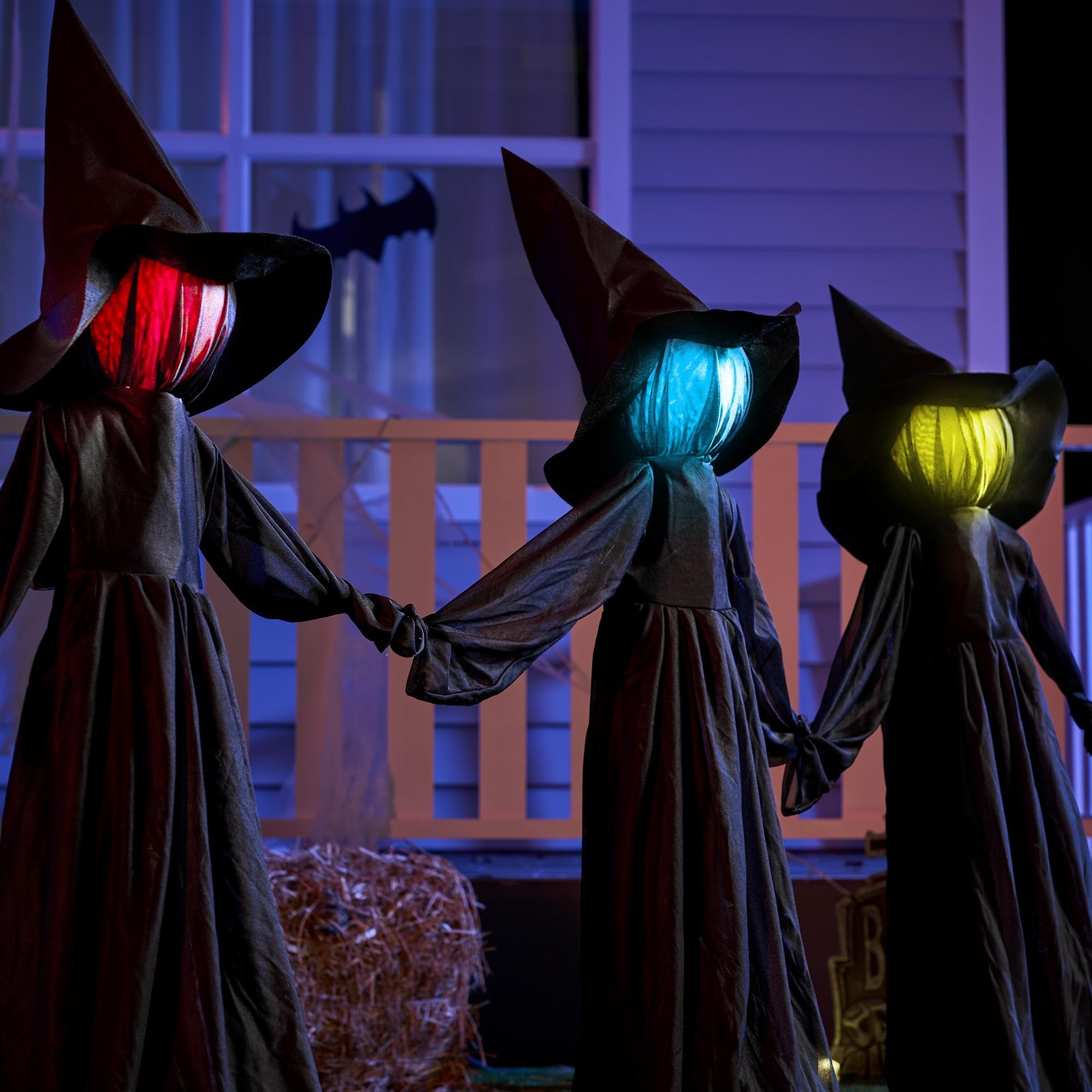 Three light-up witch yard stakes holding hands, with red, blue, and yellow glowing heads, set up on a porch at night.
