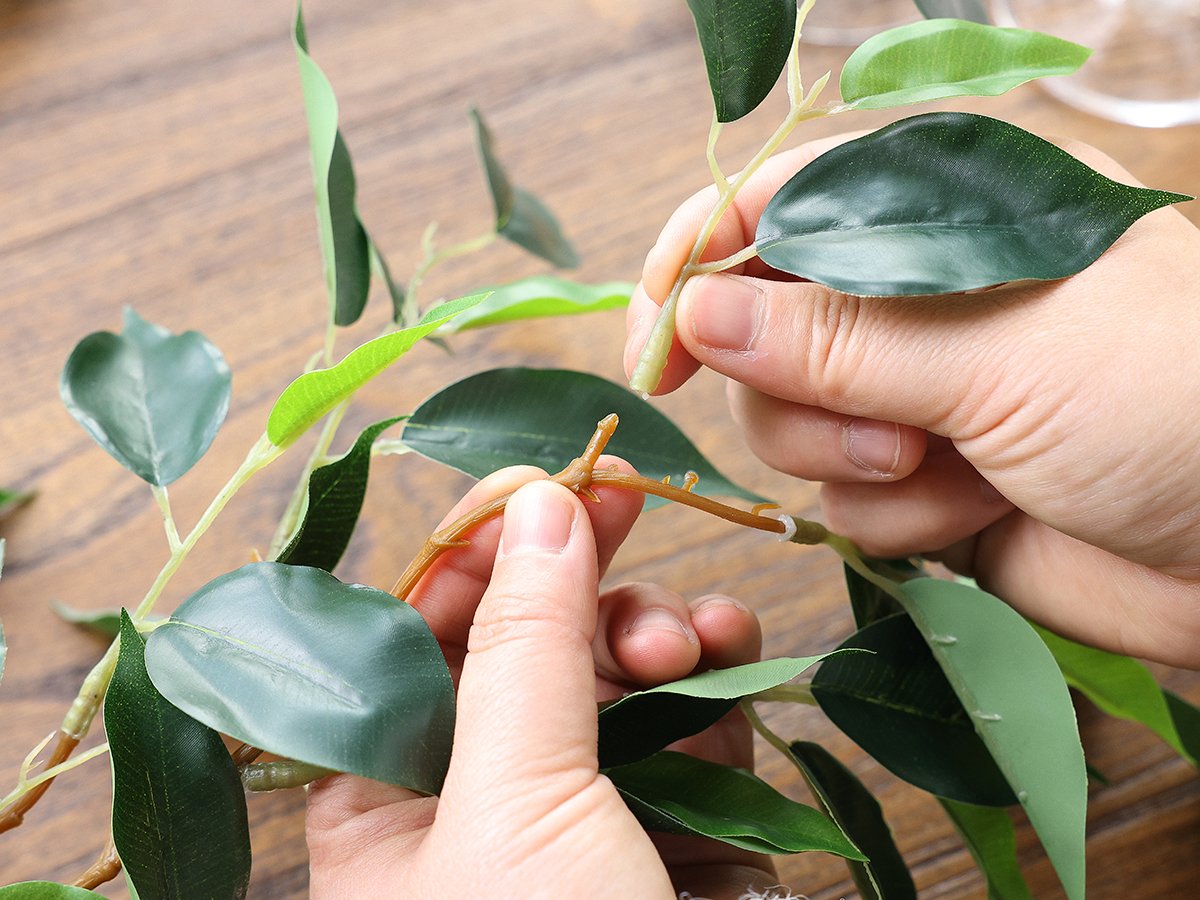 Hands assembling artificial ficus vine garland with green silk leaves and brown plastic branches on a wooden surface.