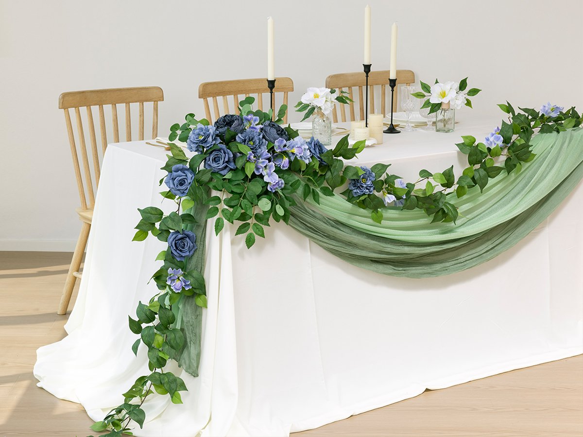 Artificial blue rose and green leaf garland draped across a white tablecloth with chairs in the background.