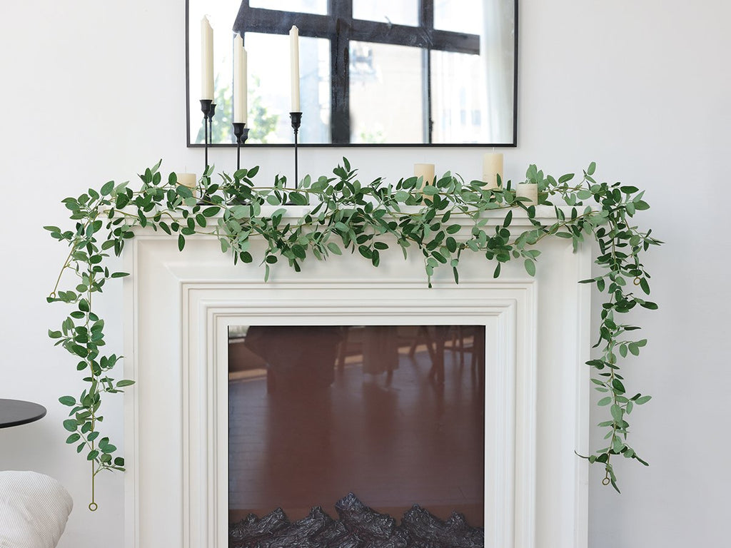 Artificial ficus vine garland with green leaves draped over a white fireplace mantel with candles and a mirror.