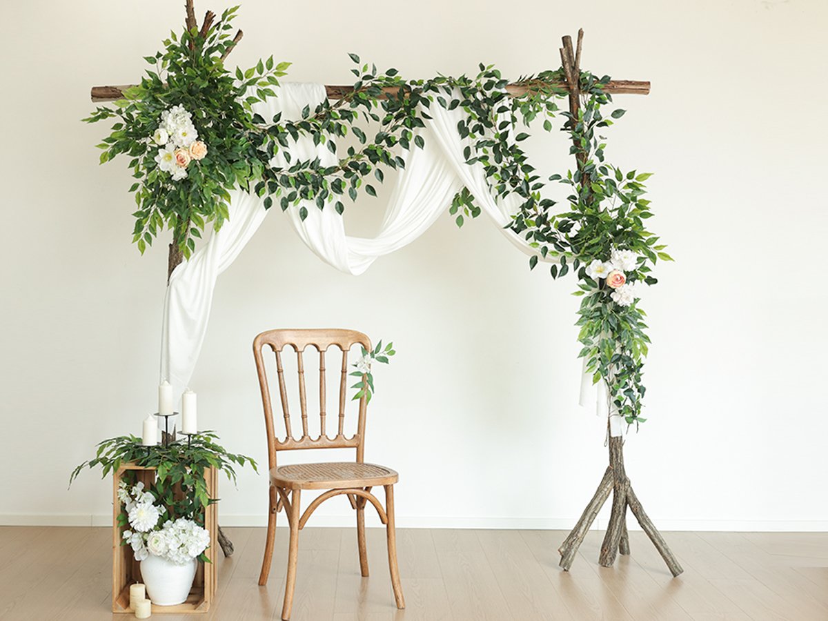 Wedding arch decorated with artificial ficus vines, white fabric, and flowers, with a chair and floral arrangement nearby.
