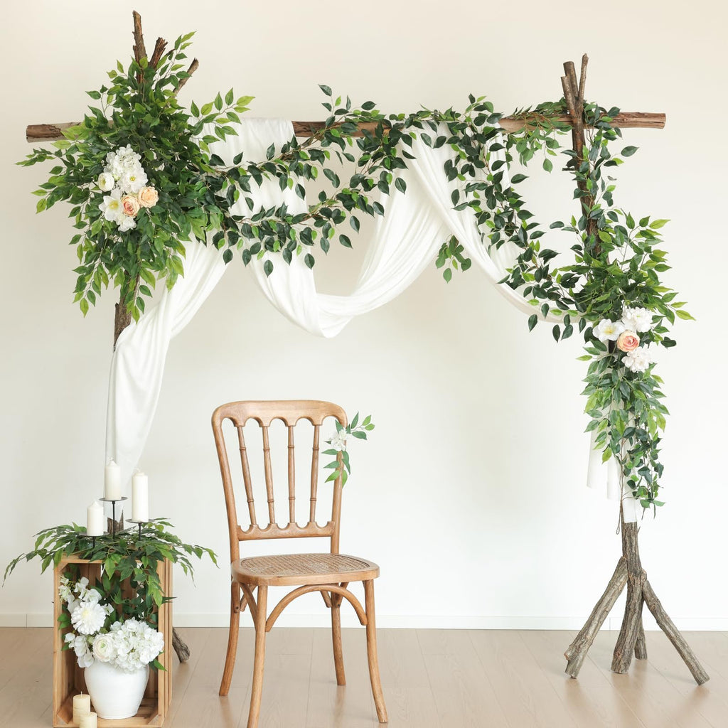 Wedding arch decorated with artificial ficus vines, white drapery, and flowers, with a wooden chair and candle display.