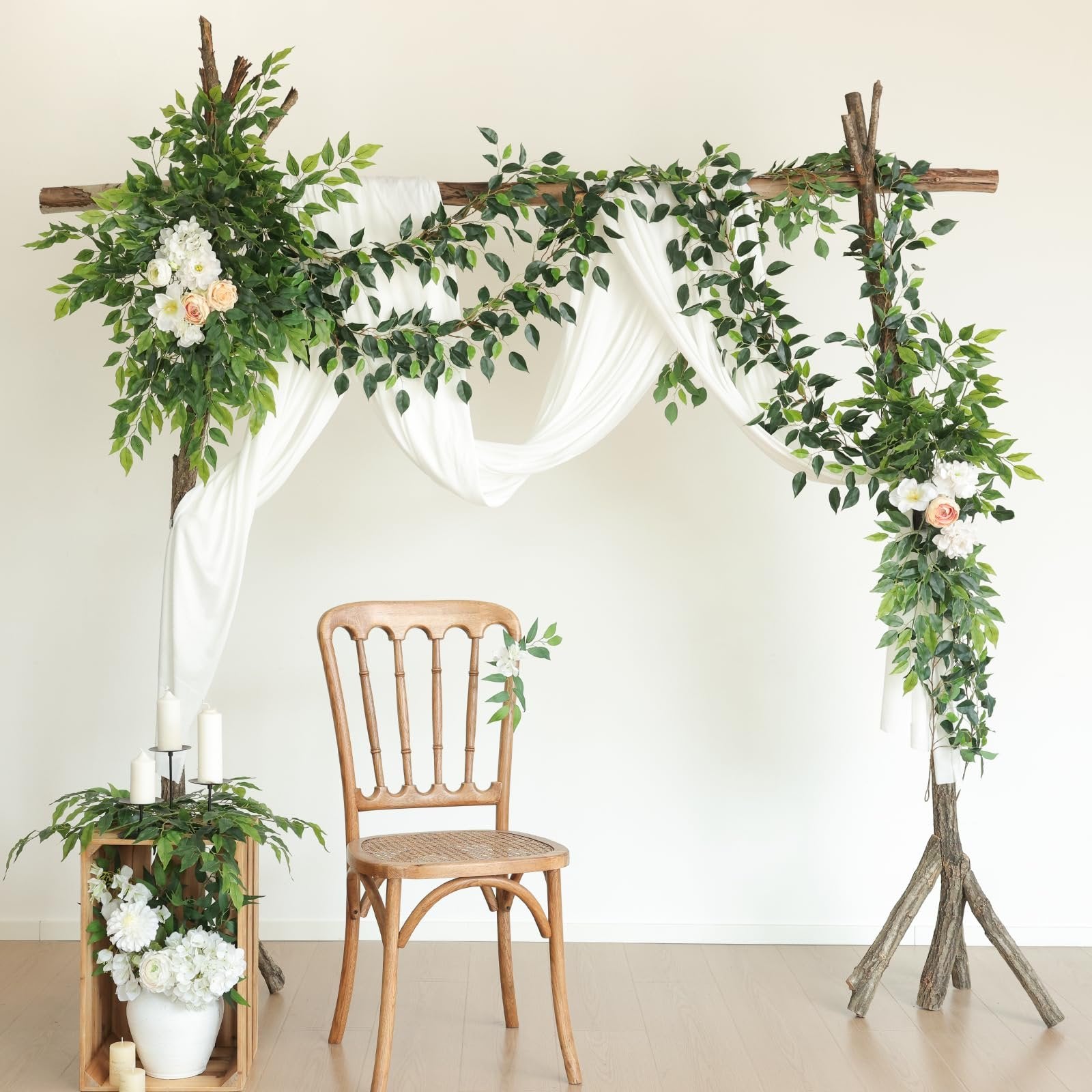 Wedding arch decorated with artificial ficus vines, white drapery, and flowers, with a wooden chair and candle display.