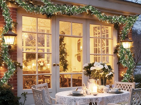 Artificial eucalyptus garland with LED lights draped over French doors and a lamp post, with a table set for dining outdoors.