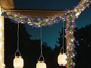 Artificial eucalyptus garland with purple flowers and LED lights draped over an overhang, with three hanging lanterns below.