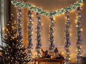 Artificial eucalyptus garland with purple flowers and LED lights draped across a wall and down pillars, with a decorated Christmas tree to the left.